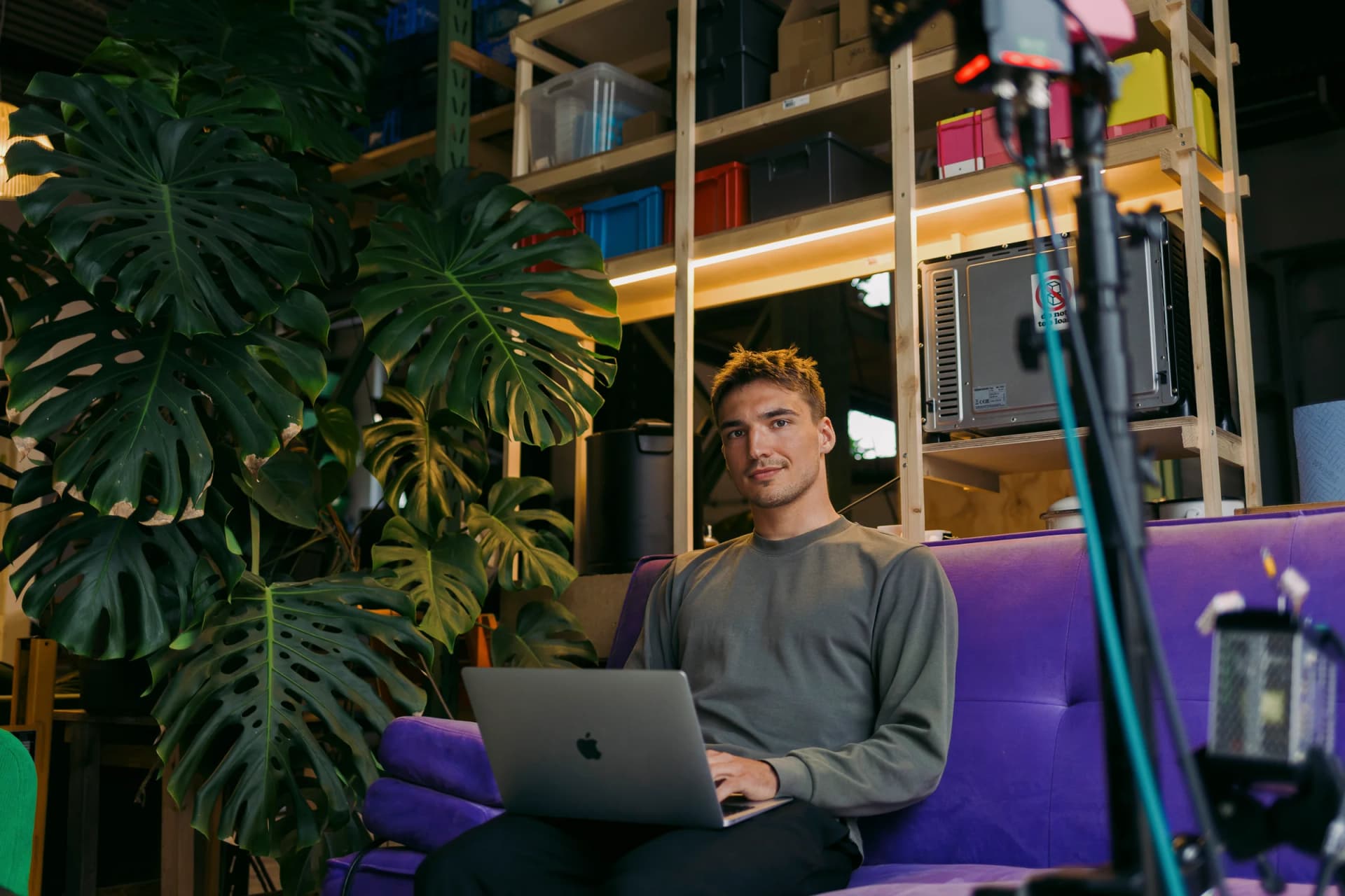 A man sits on a purple sofa in a creative studio, smiling while working on a laptop. Behind him are large monstera plants and wooden shelves with colourful storage boxes and studio equipment; a camera tripod stands in the foreground.