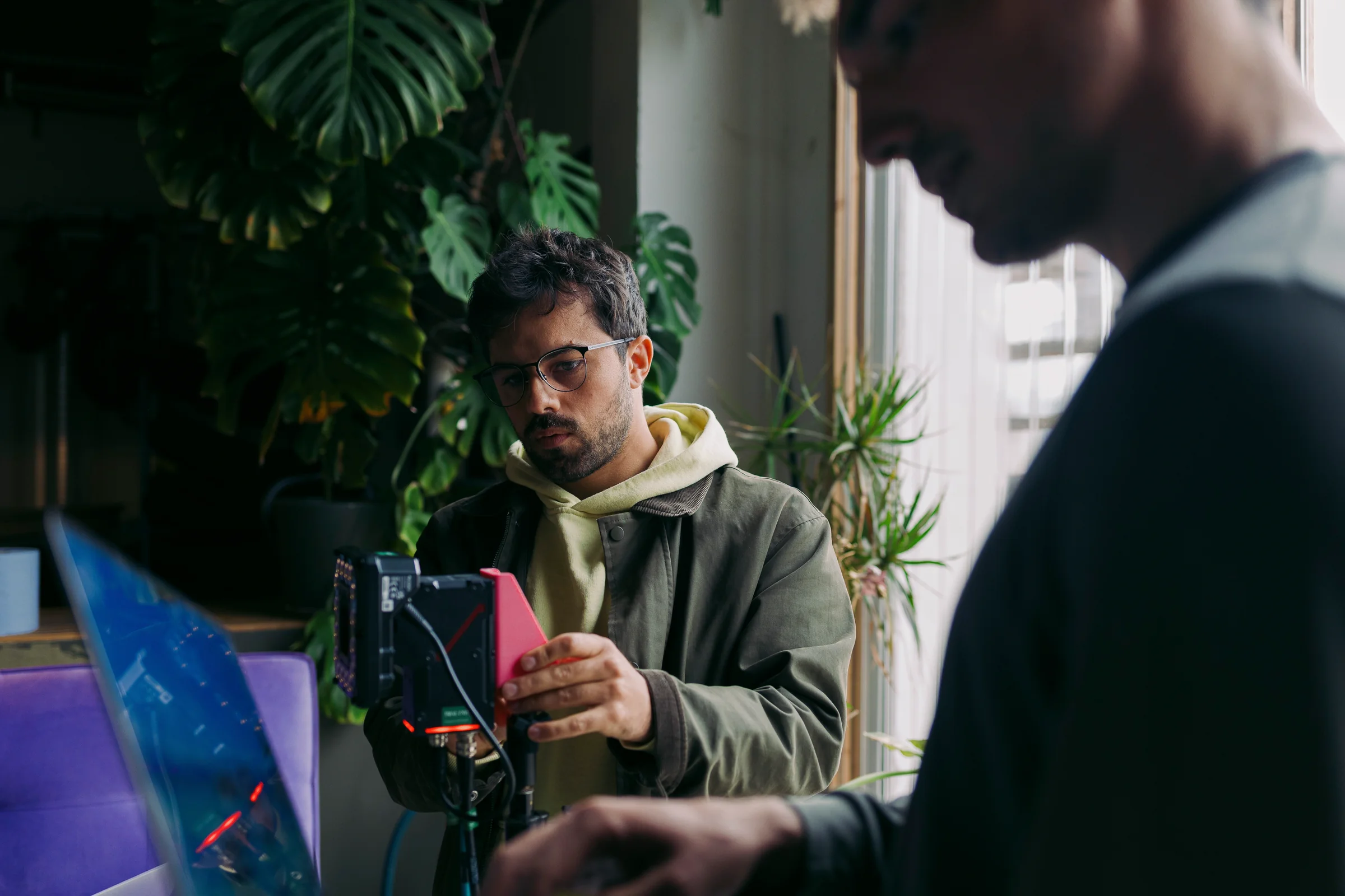 Zwei Männer arbeiten in einem Kreativstudio an einer Kamera- und Aufnahme-Ausrüstung. Einer justiert konzentriert ein Gerät; im Vordergrund ein weiterer Kollege, im Hintergrund Pflanzen und Tageslicht durch ein Fenster.