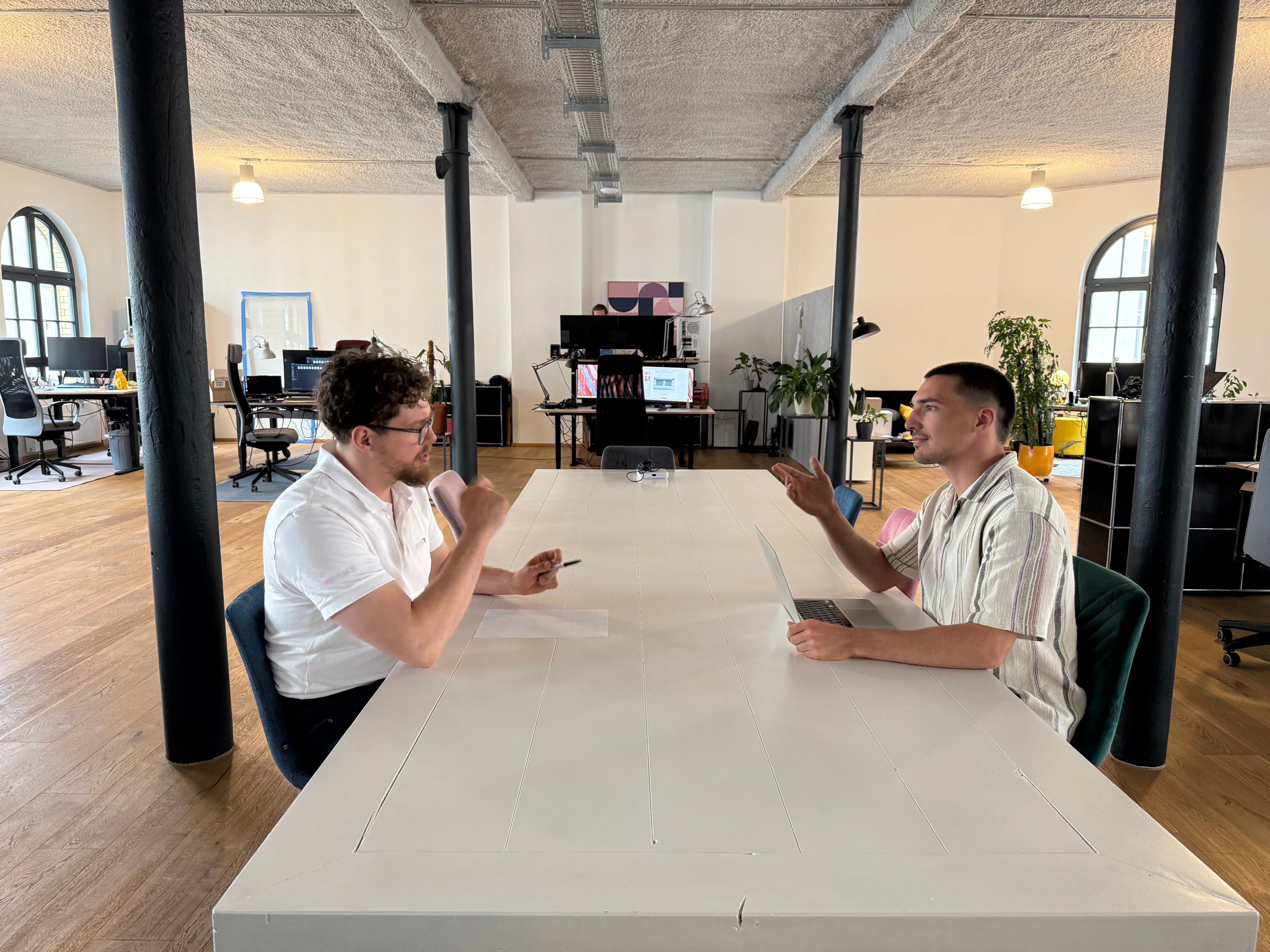 A bright, modern open-plan office with wooden floors and black support columns. Two men sit across from each other at a long white table. The man on the left wears glasses and a white polo shirt, holds a pen, and gestures as he speaks. The man on the right wears a light-striped short-sleeve shirt, holds a laptop, and responds. Behind them are desks with computer monitors, office chairs, and potted plants under exposed ceiling fixtures.