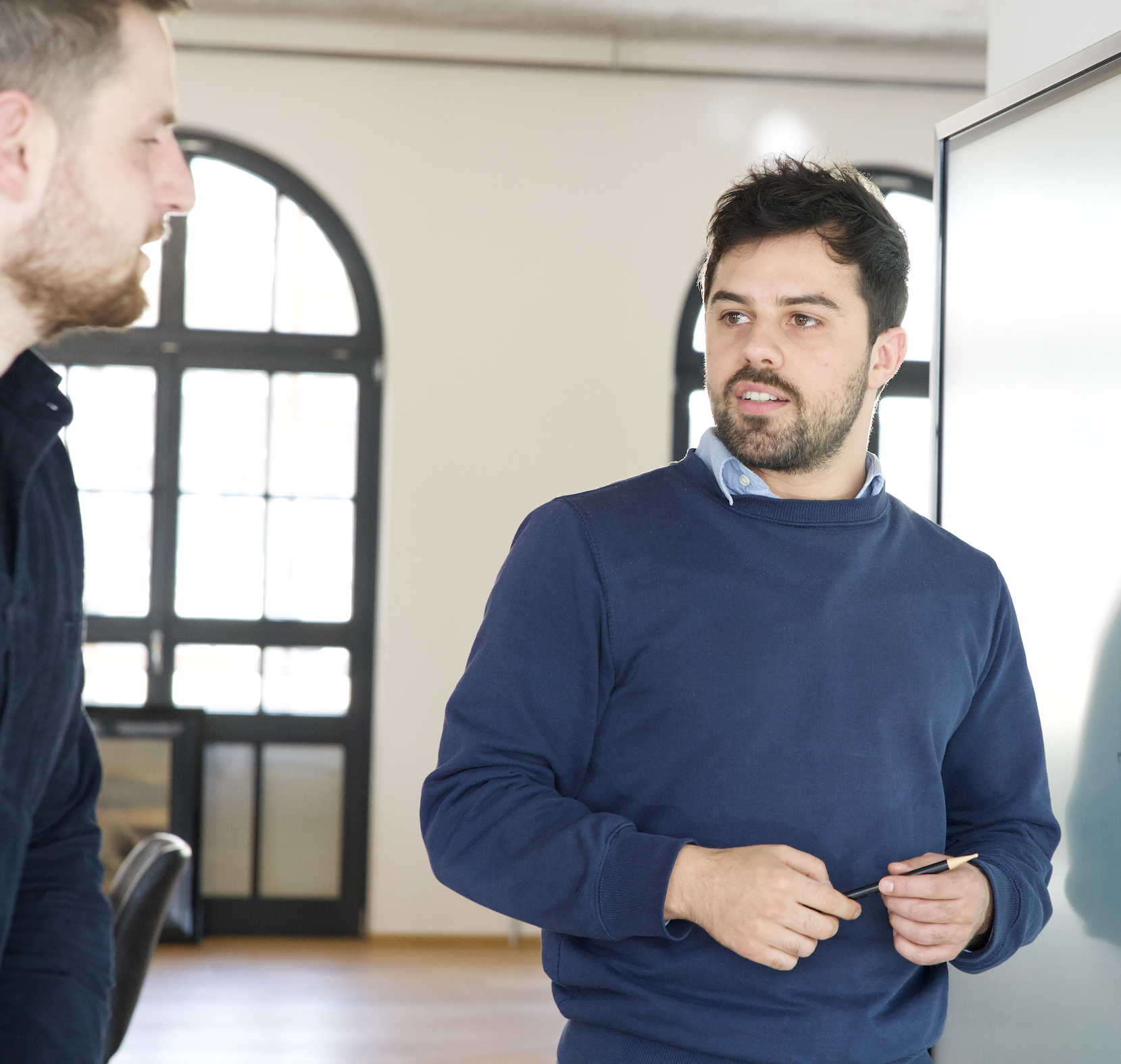 Two men are standing and talking in a bright office space. The man on the right, wearing a blue sweater and holding a pen, appears to be explaining something while facing a whiteboard or screen. The man on the left listens attentively. Large arched windows in the background let in natural light.
