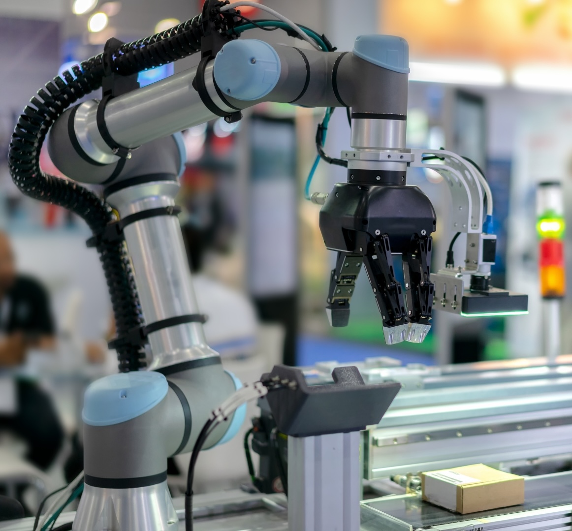 A robotic arm with a claw-like gripper is operating on an industrial assembly line. The robot appears to be picking up or placing a small cardboard box on a conveyor belt. The background shows a blurred trade show or factory setting with people and other equipment.